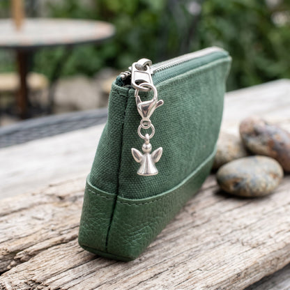 Hackberry Creek silver guardian angel charm clipped onto a green canvas and leather zipper pouch on a rustic table