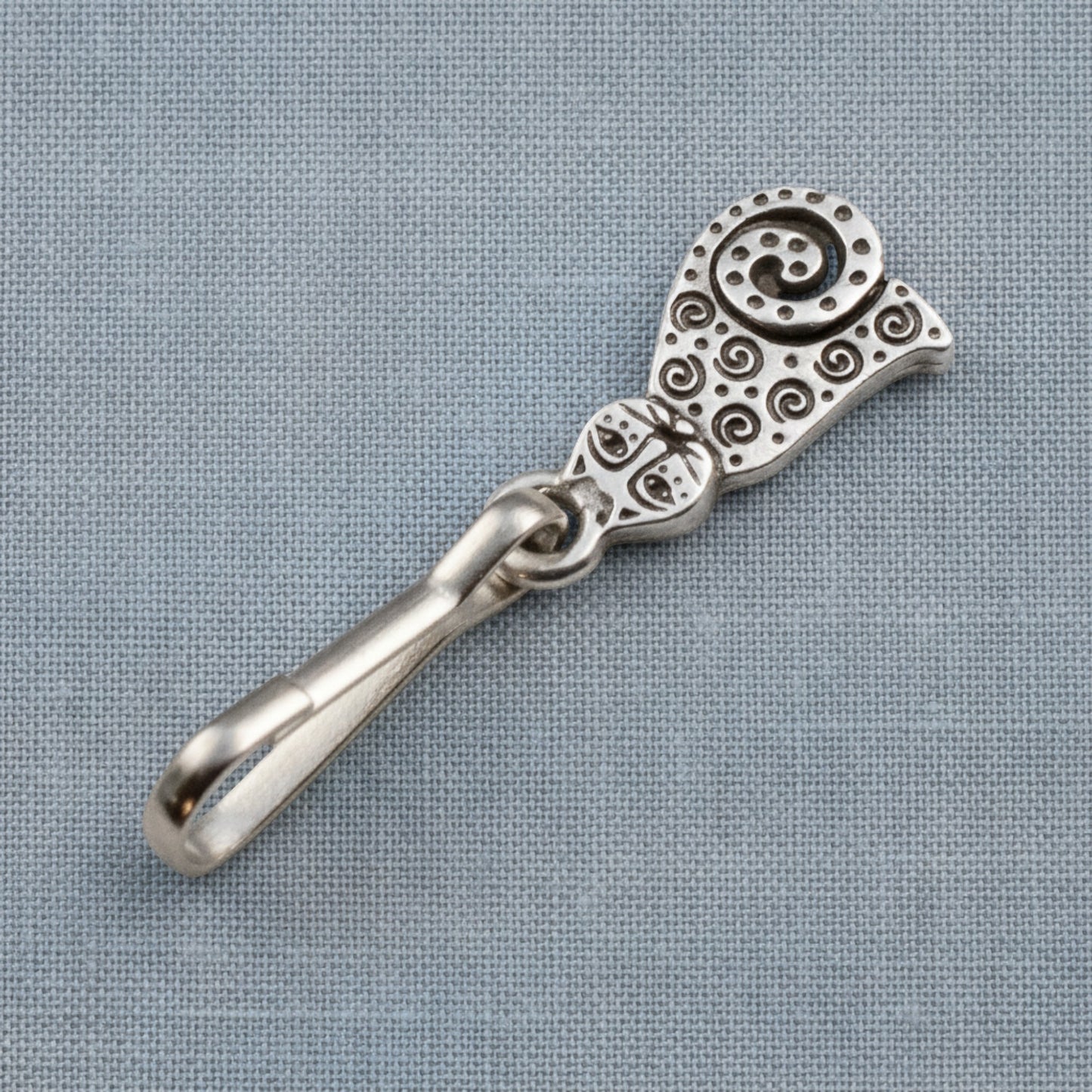 Hackberry Creek silver-plated pewter sitting cat zipper charm with intricate spiral detailing on a grey fabric background
