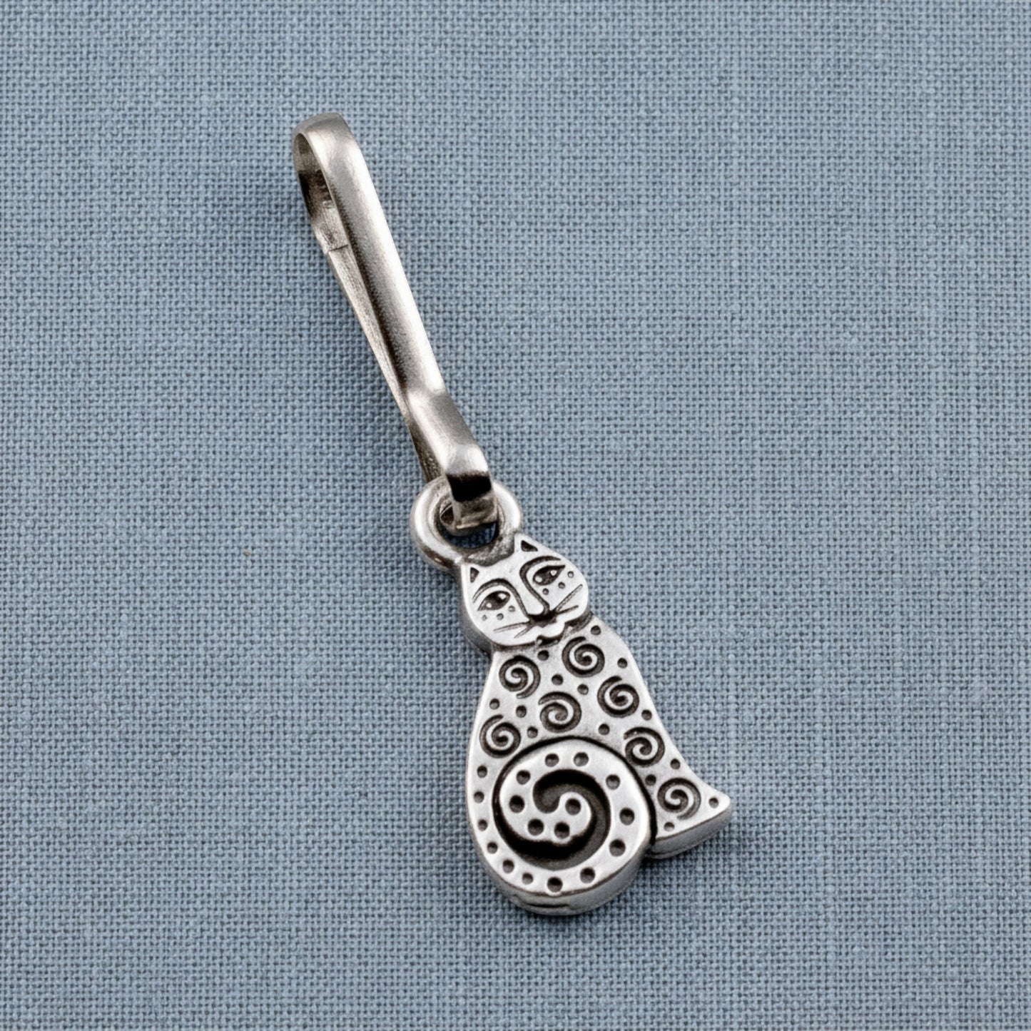 Hackberry Creek silver-plated pewter sitting cat zipper charm with intricate spiral detailing on a blue fabric background.