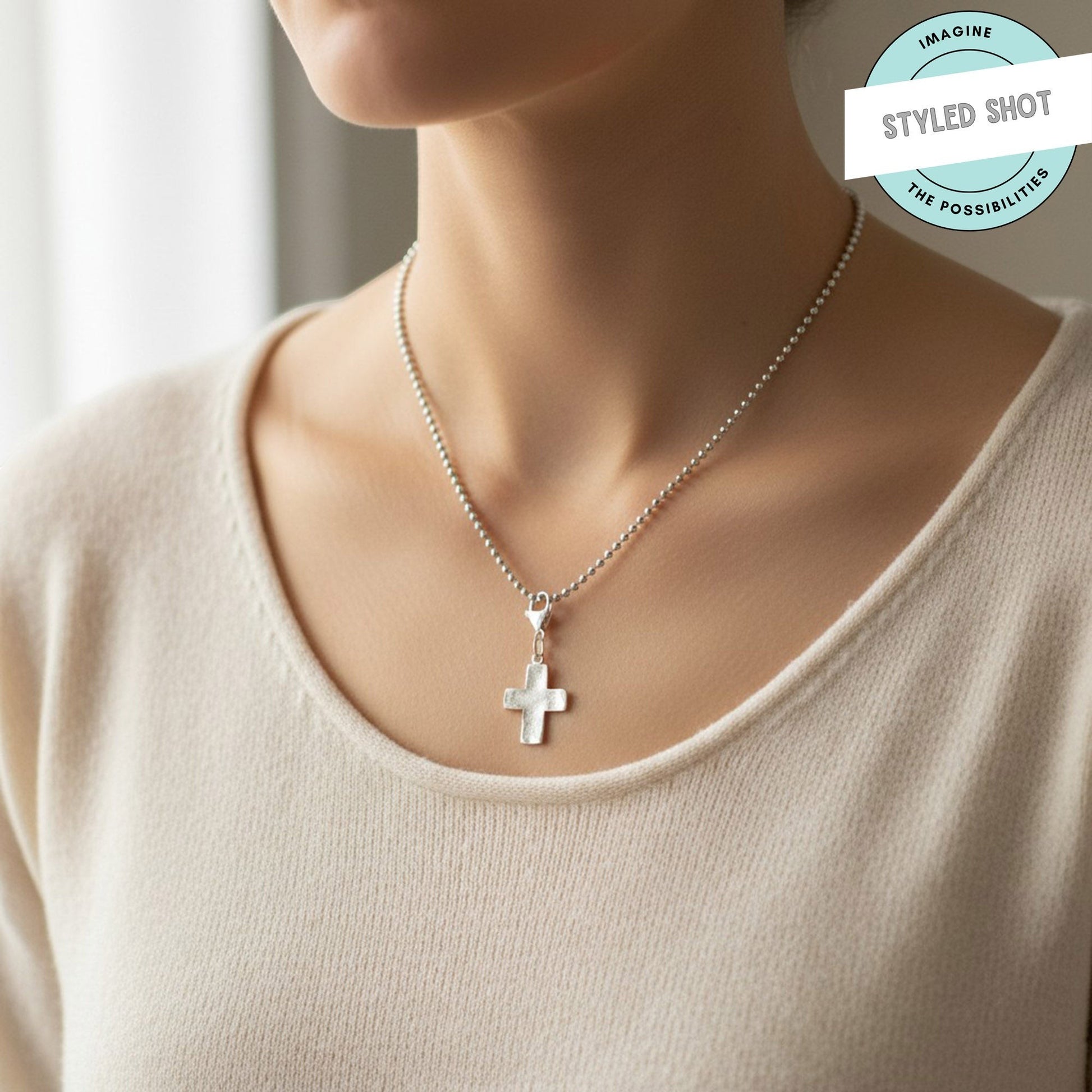 Hackberry Creek silver distressed cross charm worn as a necklace pendant on a ball chain against a cream top