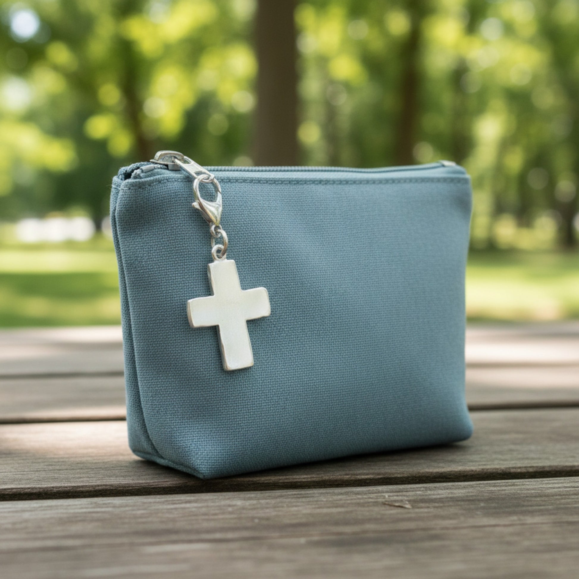 Hackberry Creek silver distressed cross charm clipped onto a blue canvas zipper pouch on a wooden table.