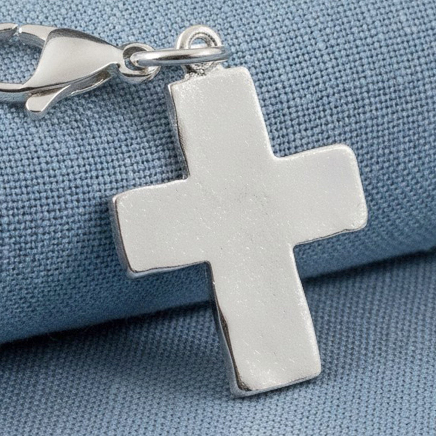 Hackberry Creek silver distressed cross charm with a stainless steel lobster clasp on a blue fabric background.