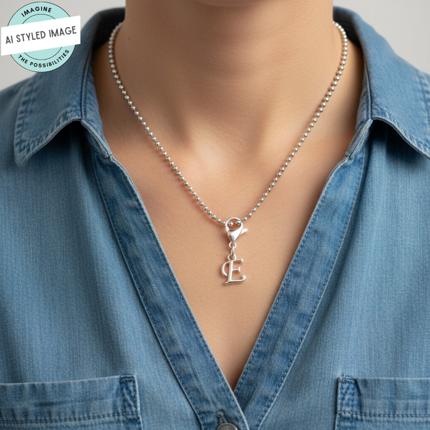 Hackberry Creek silver cursive letter E charm on a ball chain necklace worn by a person in a denim shirt.