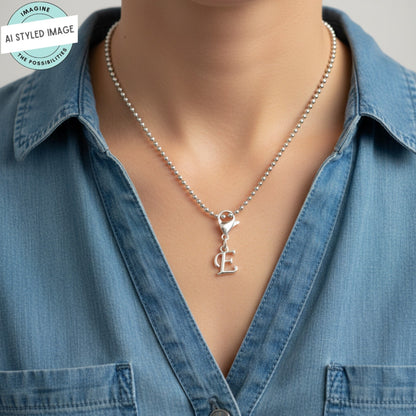 Hackberry Creek silver cursive letter E charm on a ball chain necklace worn by a person in a denim shirt.