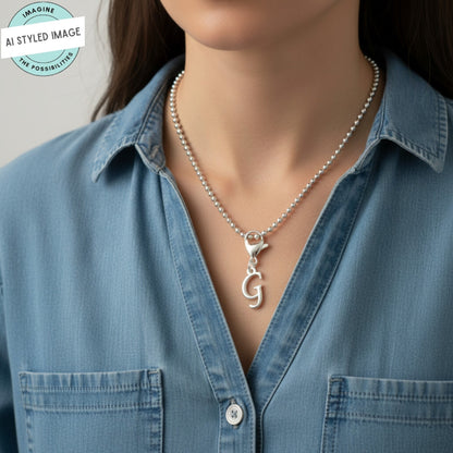 Hackberry Creek silver cursive letter G charm on a beaded necklace worn by a woman in a denim shirt.