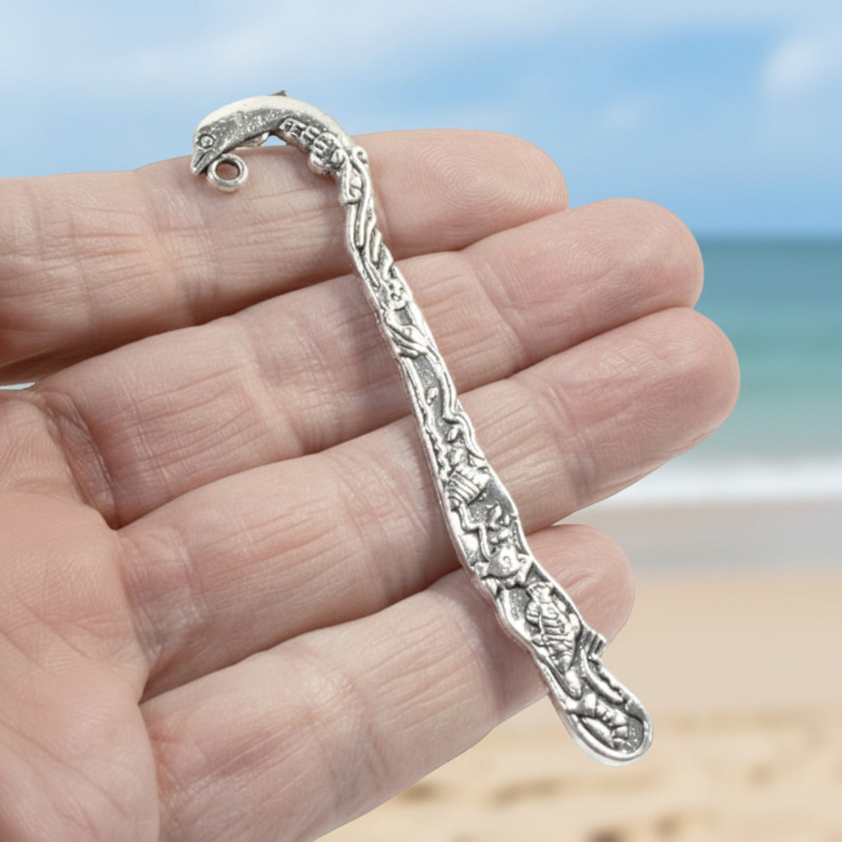 Hackberry Creek antique silver dolphin bookmark with beach-themed engraving held in hand against a blurred beach background