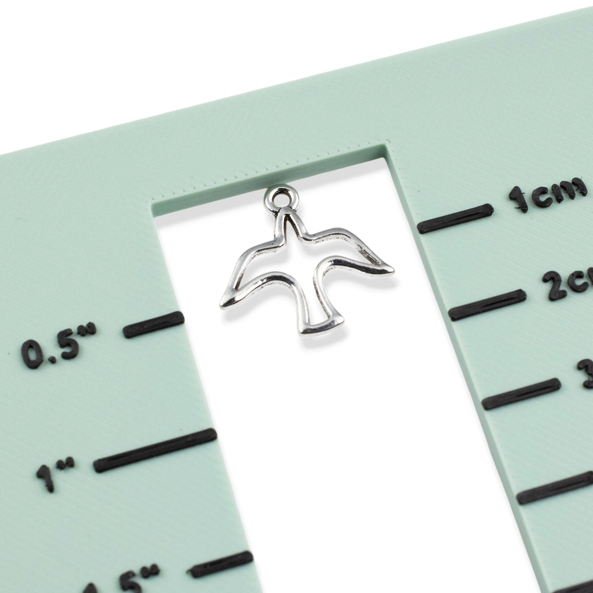 Hackberry Creek silver peace dove charm with open silhouette design on a green measuring tool for jewelry making.