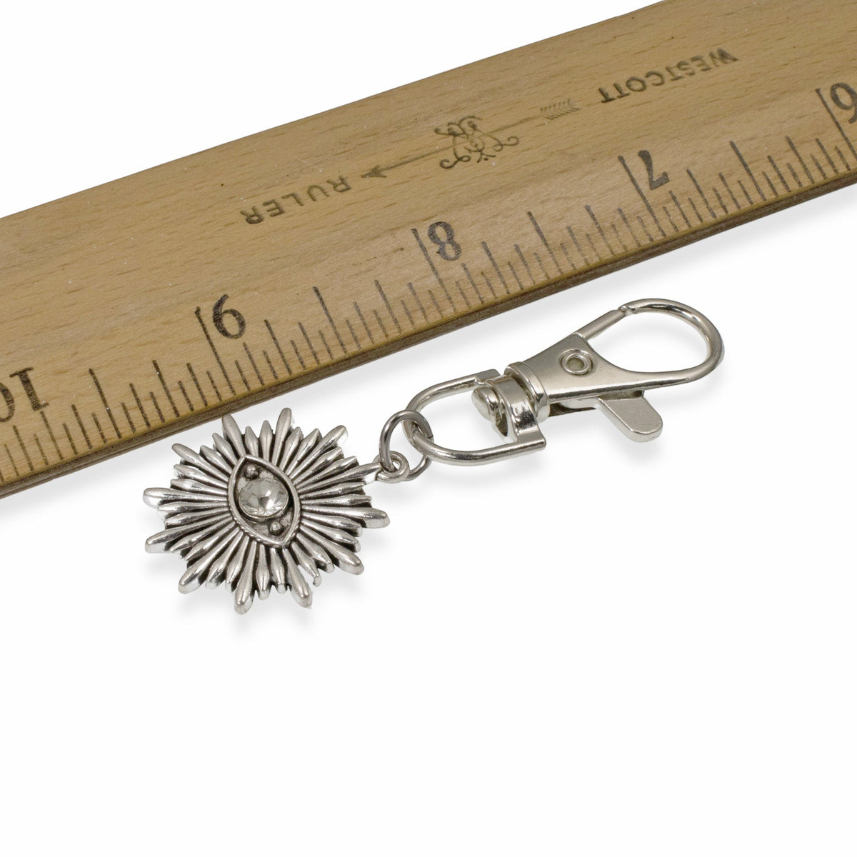 Hackberry Creek silver metal alloy evil eye sunburst key fob with a clear rhinestone accent next to a wooden ruler.