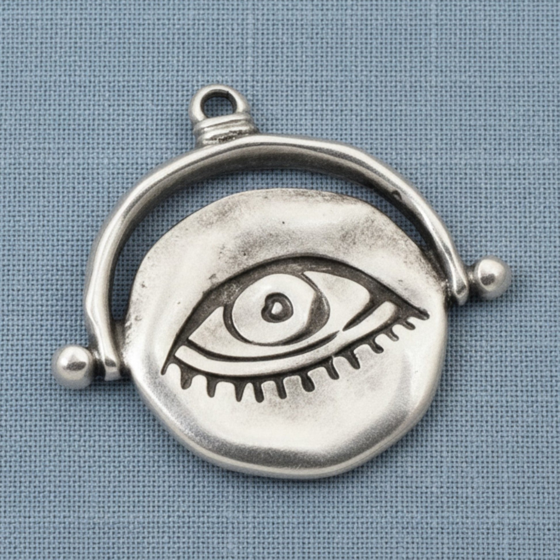 Hackberry Creek silver plated evil eye pendant with a hand-formed organic texture on a blue fabric background.