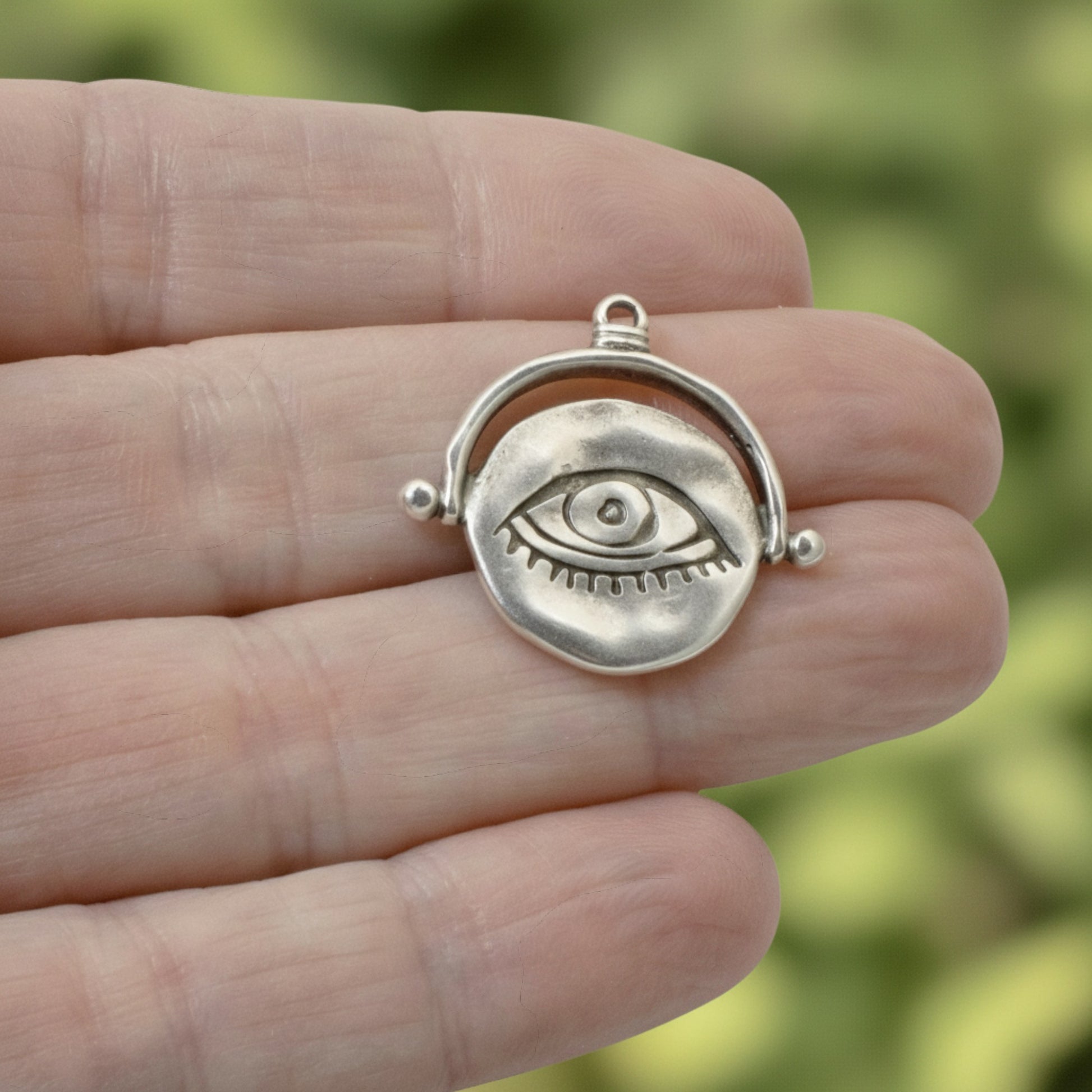 Hackberry Creek silver plated evil eye pendant with hand-formed texture held in a hand against a blurred green background