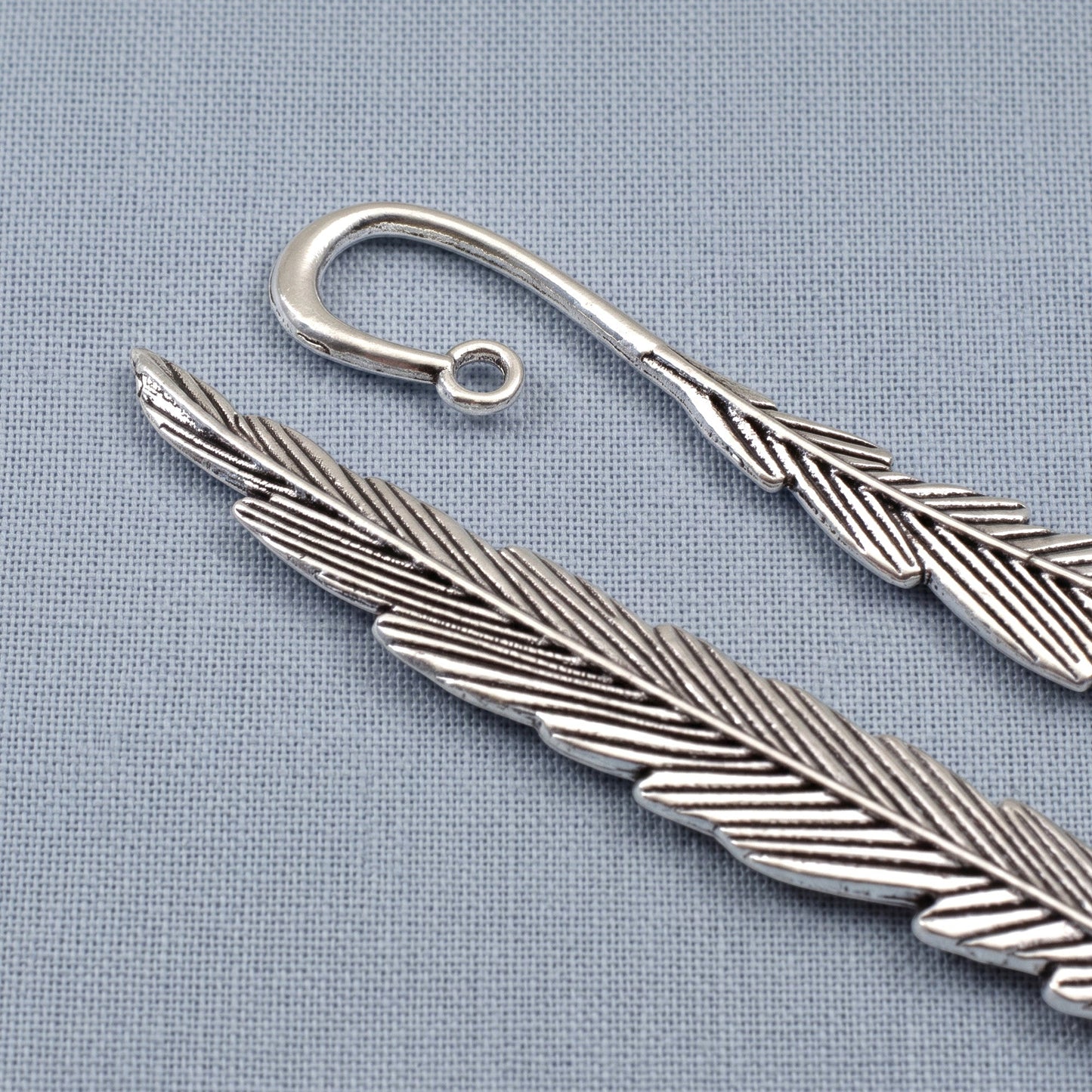 Hackberry Creek antique silver feather bookmark bases with etched detail and a charm loop on a blue fabric background.
