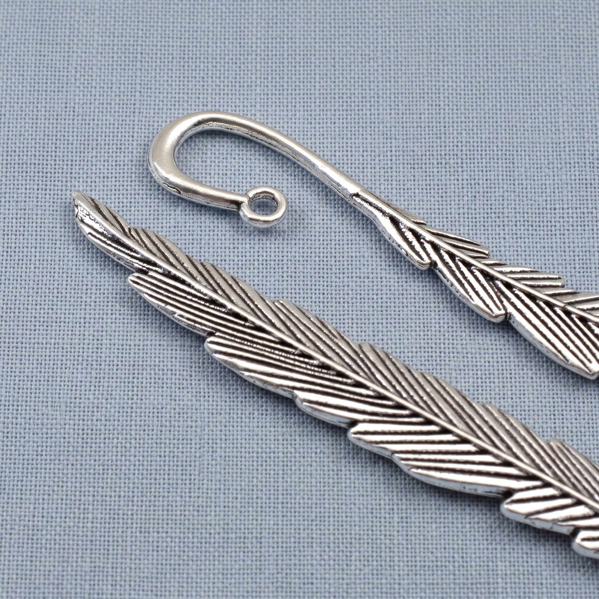 Hackberry Creek antique silver feather bookmark bases with etched detail and a charm loop on a blue fabric background.