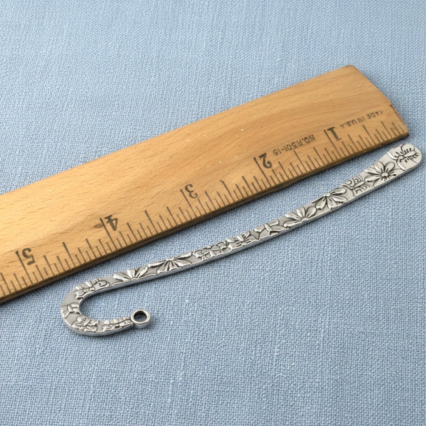 Hackberry Creek silver zinc alloy hook bookmark with raised floral and butterfly pattern next to a wooden ruler.