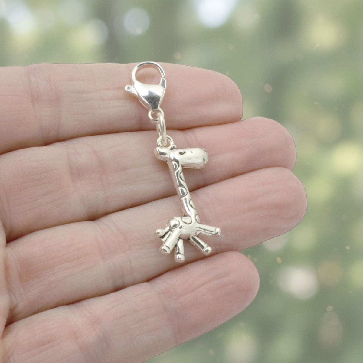 Hackberry Creek silver giraffe charm with a stainless steel lobster clasp held in a hand against a blurred background.