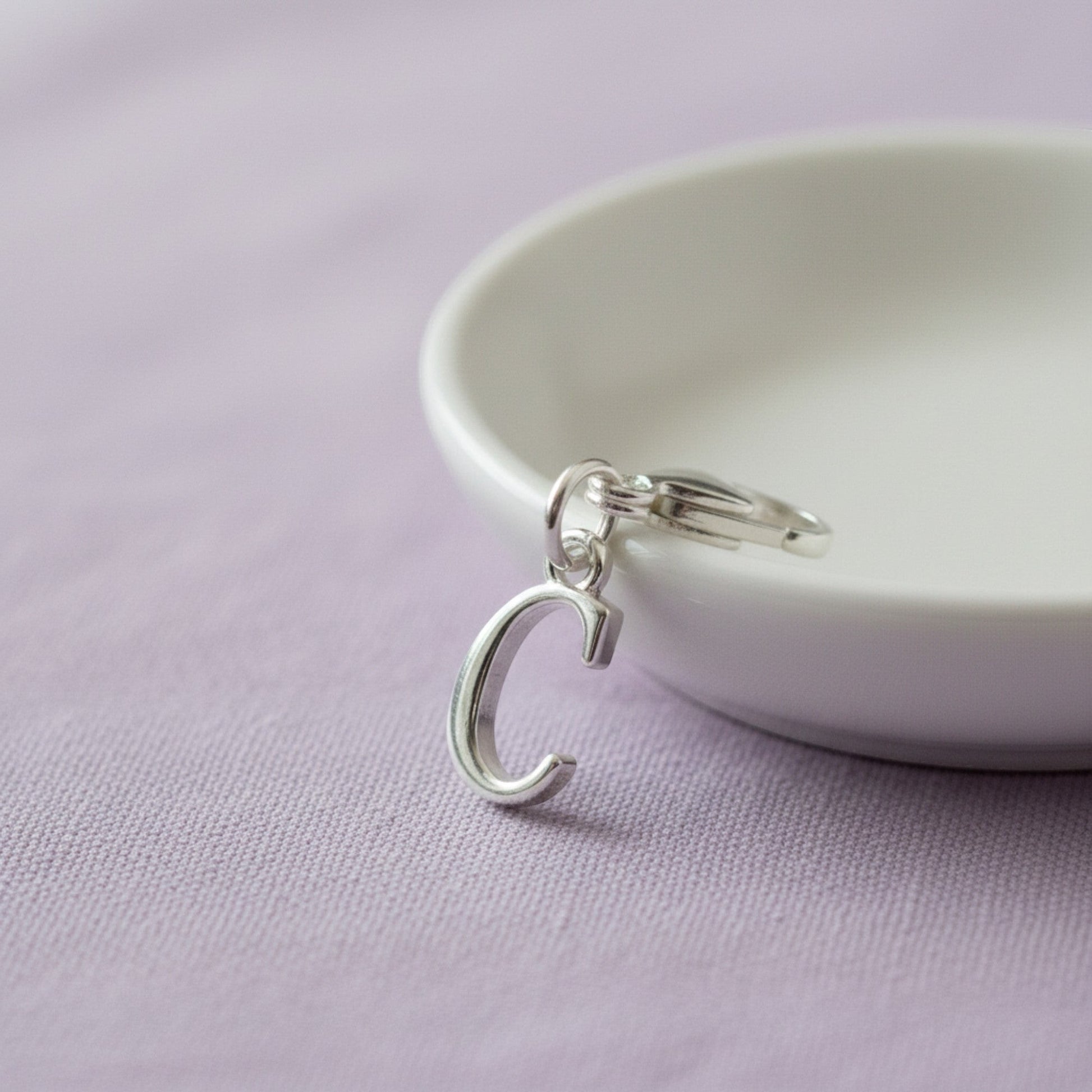 Hackberry Creek silver cursive letter C charm with a stainless steel lobster clasp on a white dish.