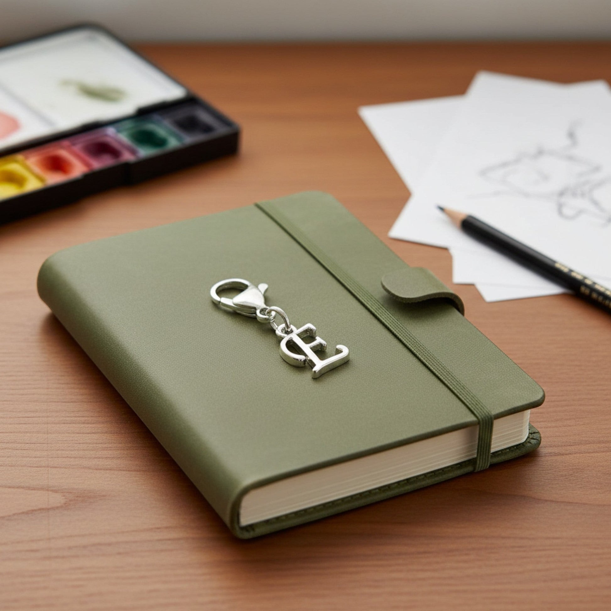 Hackberry Creek silver cursive letter E charm with lobster clasp clipped onto a green journal on a wooden desk.
