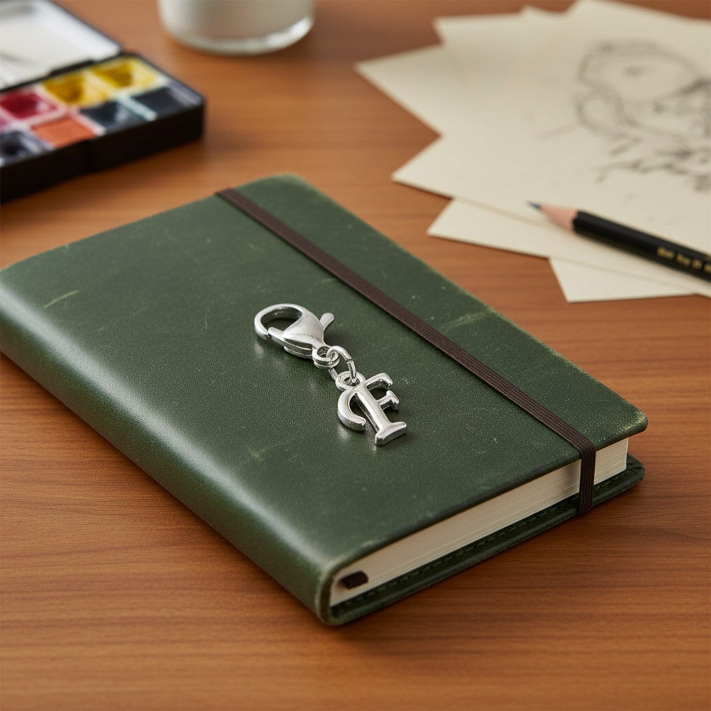 Hackberry Creek silver cursive letter F charm with lobster clasp clipped onto a green leather journal cover.