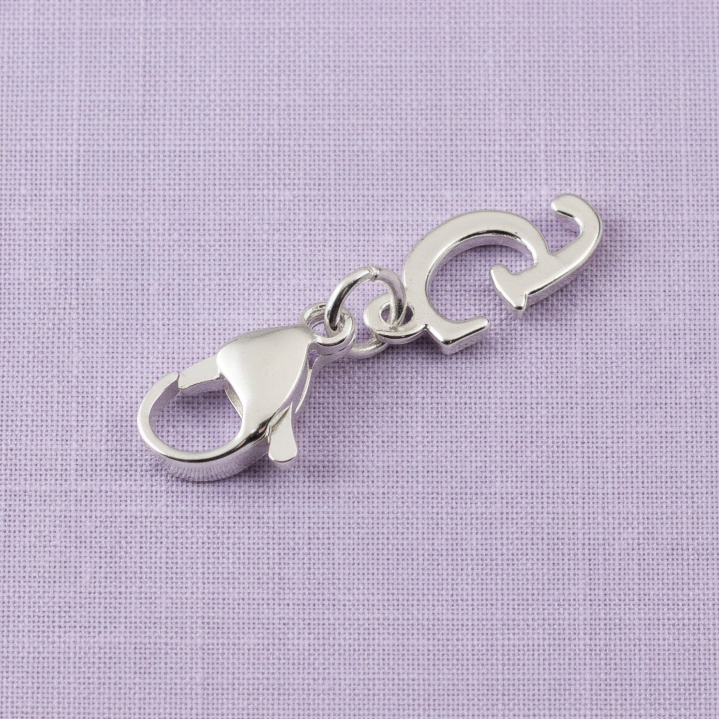 Hackberry Creek silver cursive letter G charm with a stainless steel lobster clasp on a light purple fabric background.
