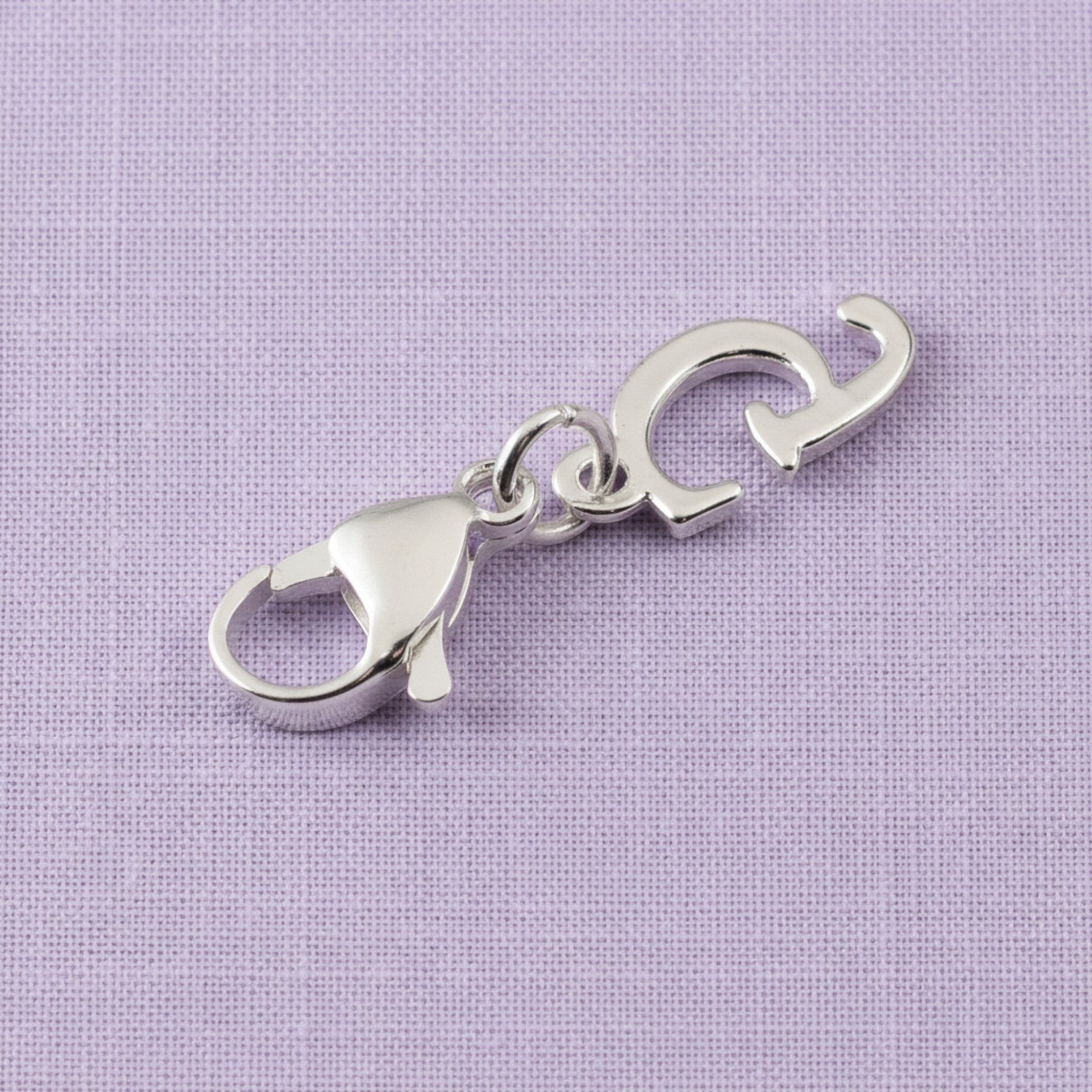 Hackberry Creek silver cursive letter G charm with a stainless steel lobster clasp on a light purple fabric background.