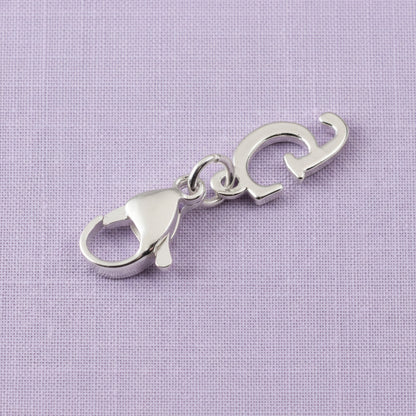 Hackberry Creek silver cursive letter G charm with a stainless steel lobster clasp on a light purple fabric background.