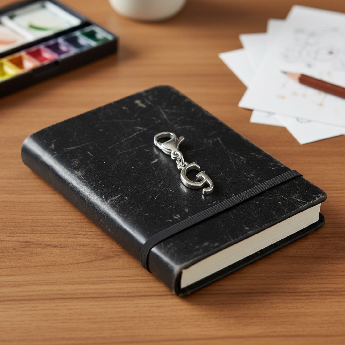 Hackberry Creek silver cursive letter G charm with lobster clasp clipped to a black journal on a wooden desk.