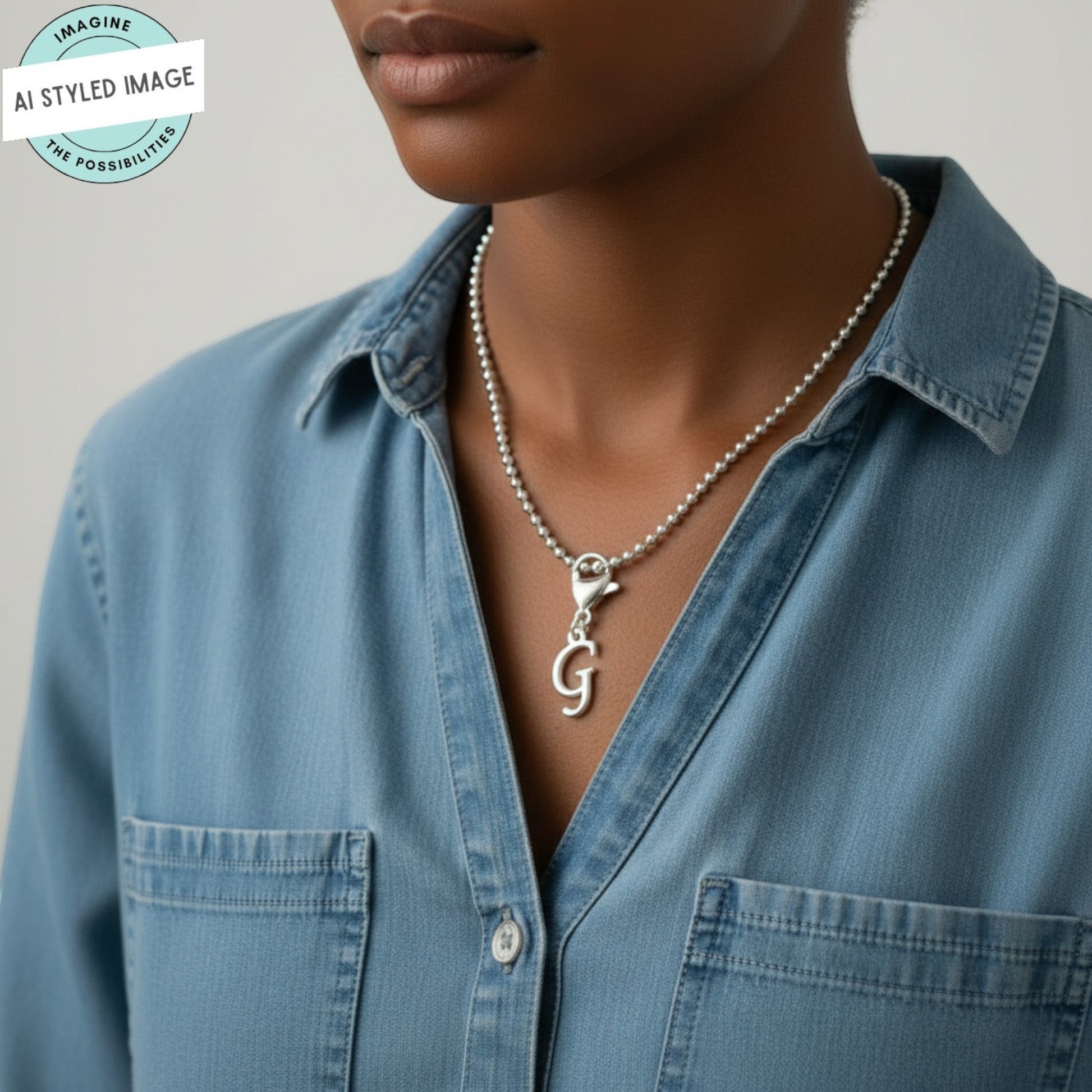 Hackberry Creek silver cursive letter G charm on a beaded necklace worn by a woman in a blue denim shirt.