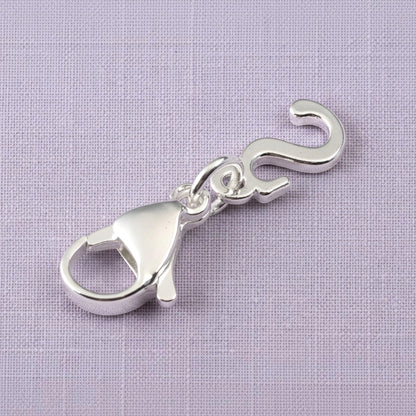 Hackberry Creek silver cursive letter S charm with a stainless steel lobster clasp on a textured purple background.