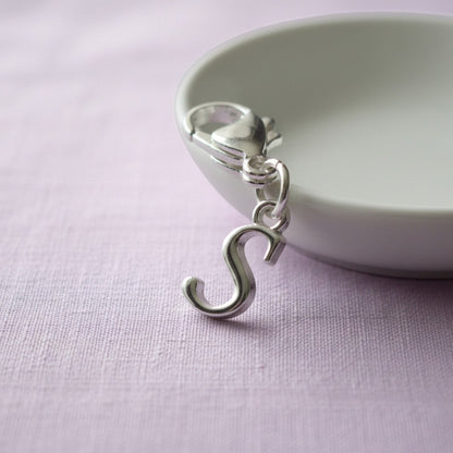 Hackberry Creek silver cursive letter S charm with a stainless steel lobster clasp resting on a small white dish.