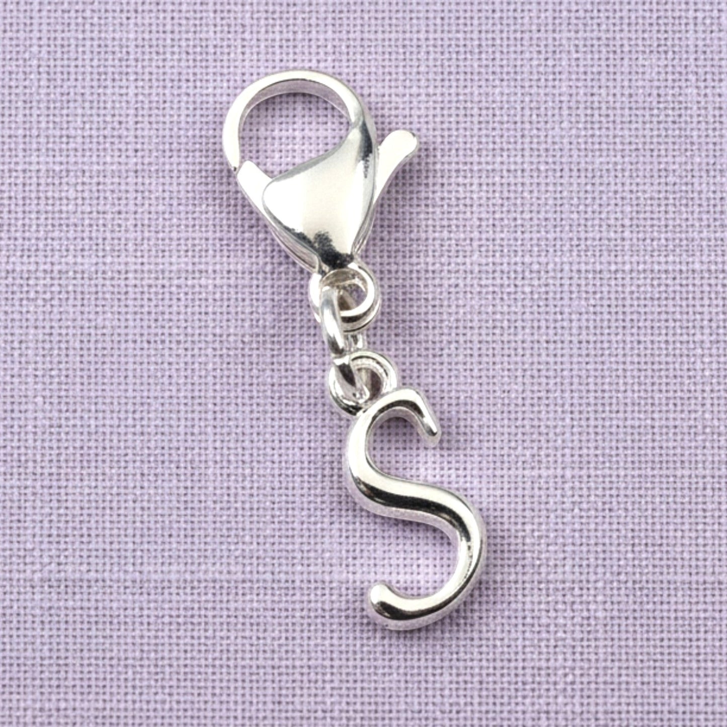 Hackberry Creek silver cursive letter S charm with a stainless steel lobster clasp on a light purple fabric background.