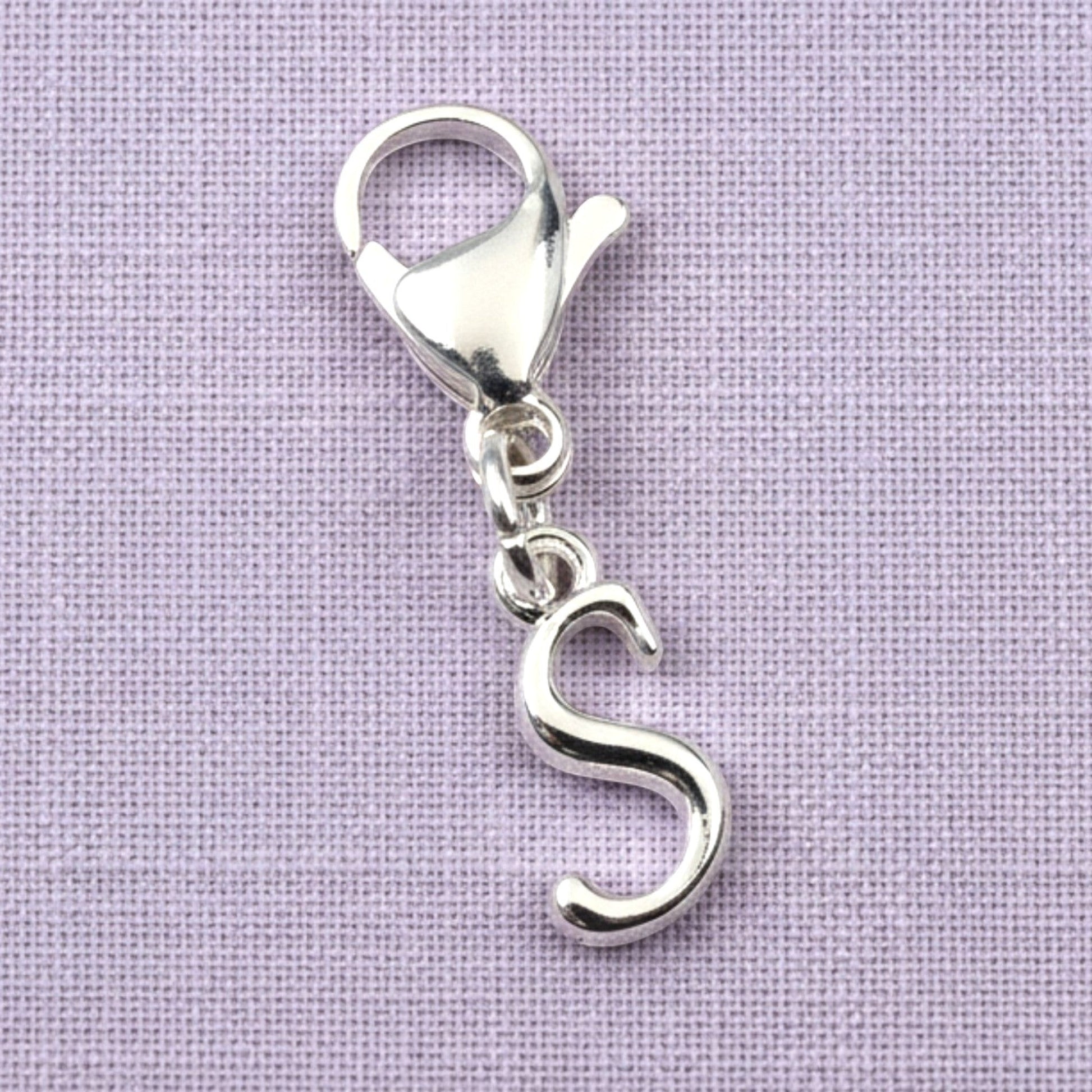 Hackberry Creek silver cursive letter S charm with a stainless steel lobster clasp on a light purple fabric background.