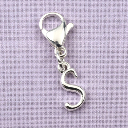 Hackberry Creek silver cursive letter S charm with a stainless steel lobster clasp on a light purple fabric background.