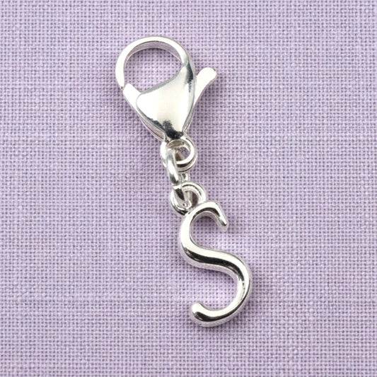 Hackberry Creek silver cursive letter S charm with a stainless steel lobster clasp on a light purple fabric background.