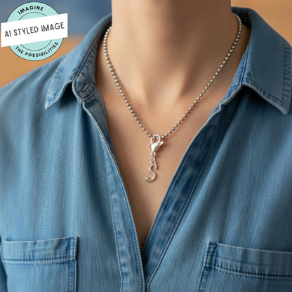 Hackberry Creek silver cursive letter S charm on a ball chain necklace worn by a person in a denim shirt.