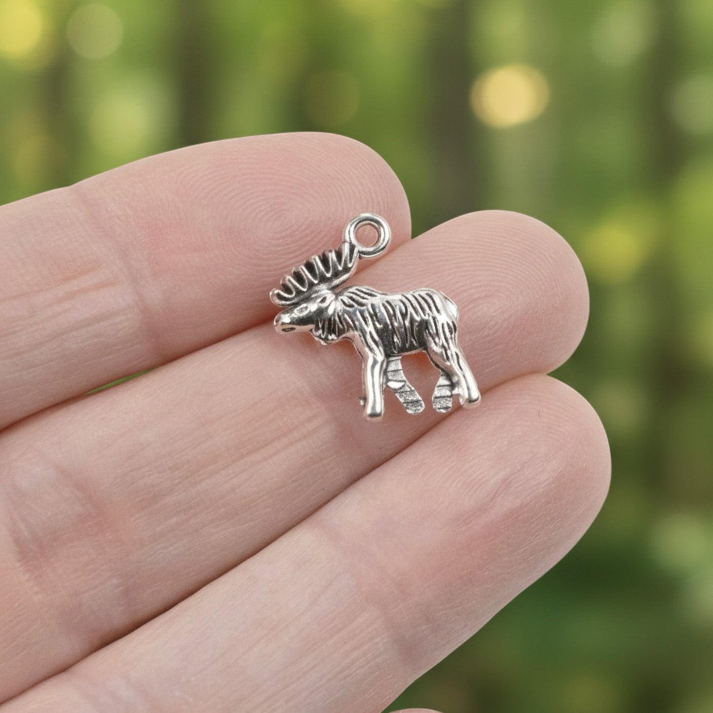 Hackberry Creek silver moose charm held between fingers against a blurred forest background.