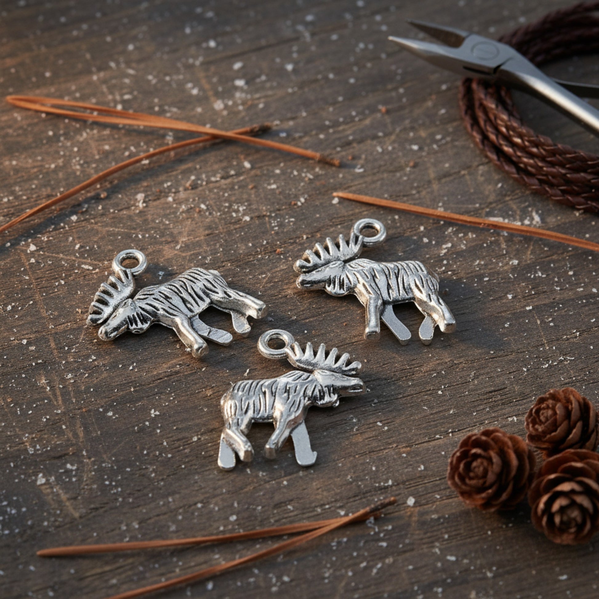 Hackberry Creek silver moose charms with detailed walking design on a rustic wooden surface with pine needles.