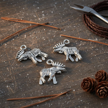 Hackberry Creek silver moose charms with detailed walking design on a rustic wooden surface with pine needles.