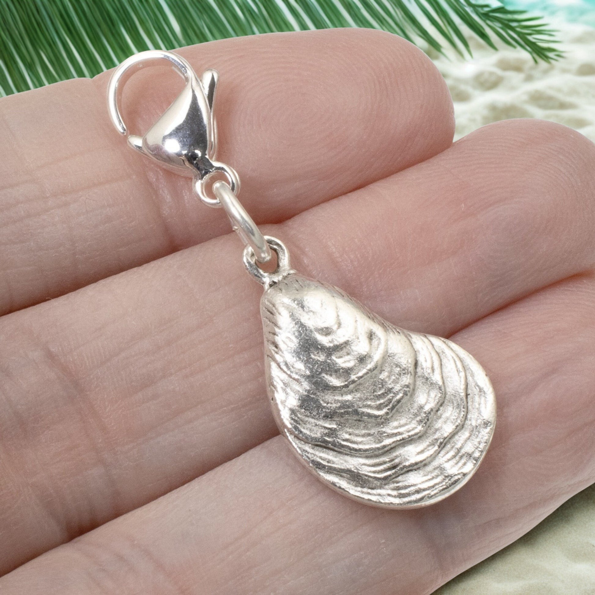 Hackberry Creek silver-plated oyster shell charm with lobster clasp held in a hand against a beach-themed background.