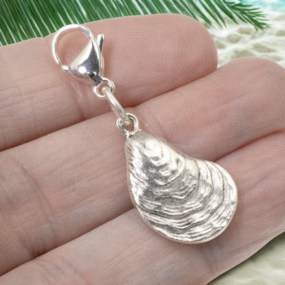 Hackberry Creek silver-plated oyster shell charm with lobster clasp held in a hand against a beach-themed background.