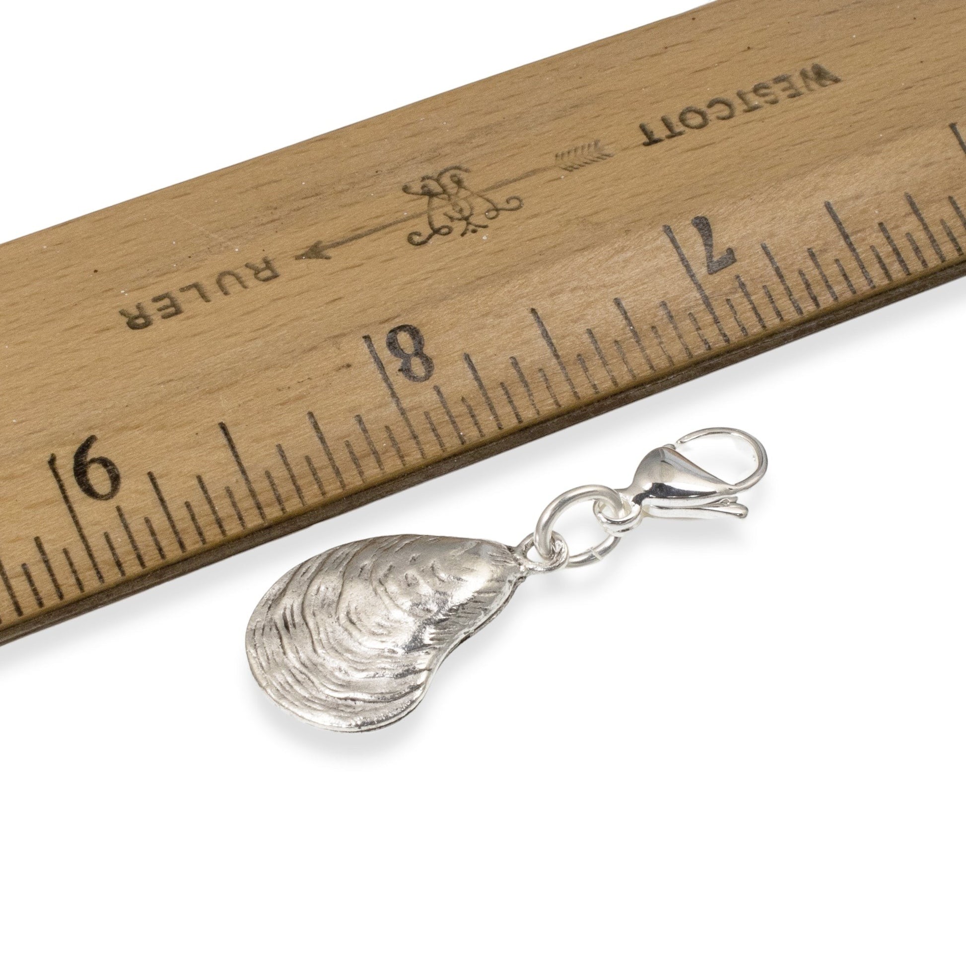 Hackberry Creek silver oyster shell charm with lobster clasp next to a wooden ruler for scale on a white background.