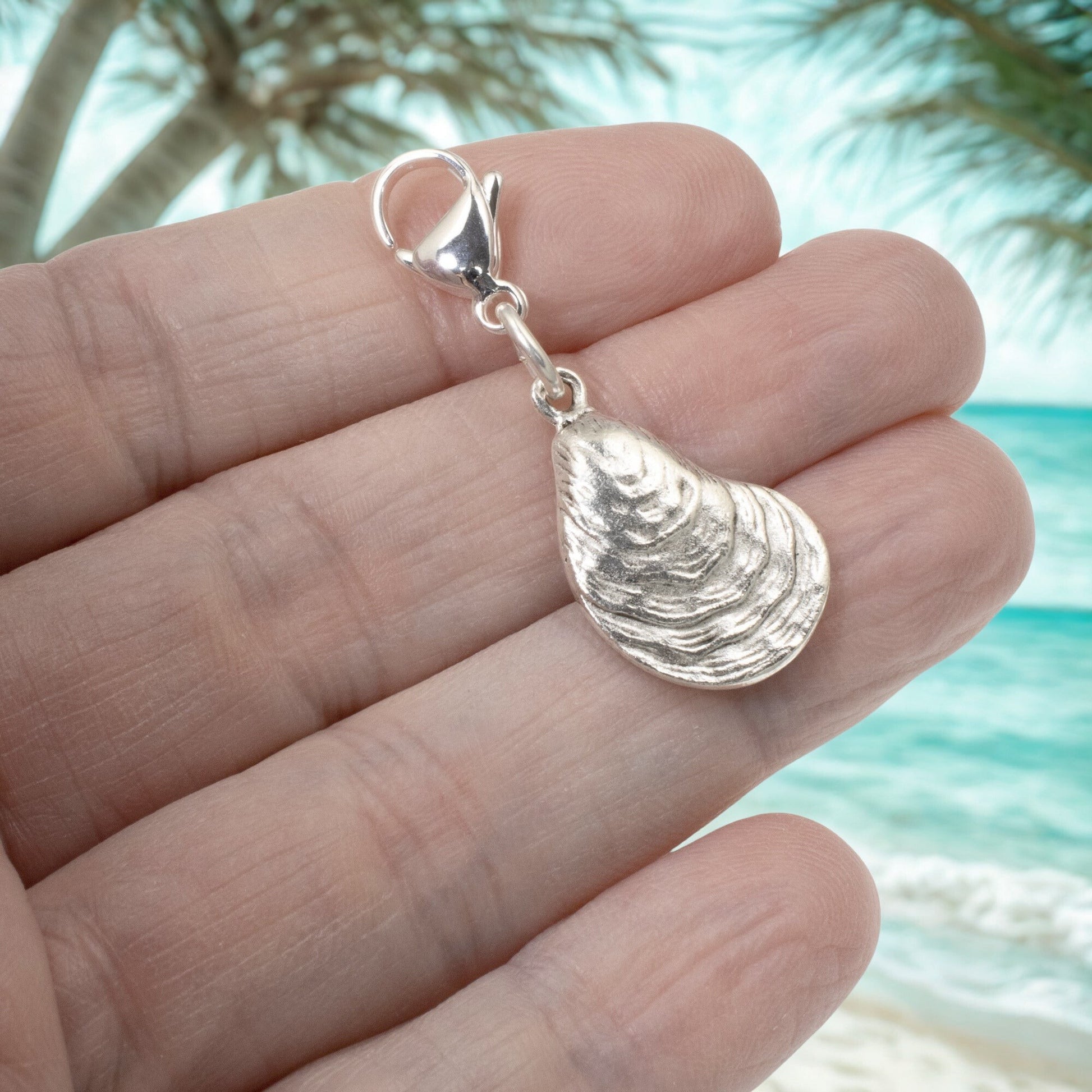 Hackberry Creek silver-plated oyster shell charm with lobster clasp held in hand against a tropical beach background.