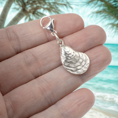 Hackberry Creek silver-plated oyster shell charm with lobster clasp held in hand against a tropical beach background.