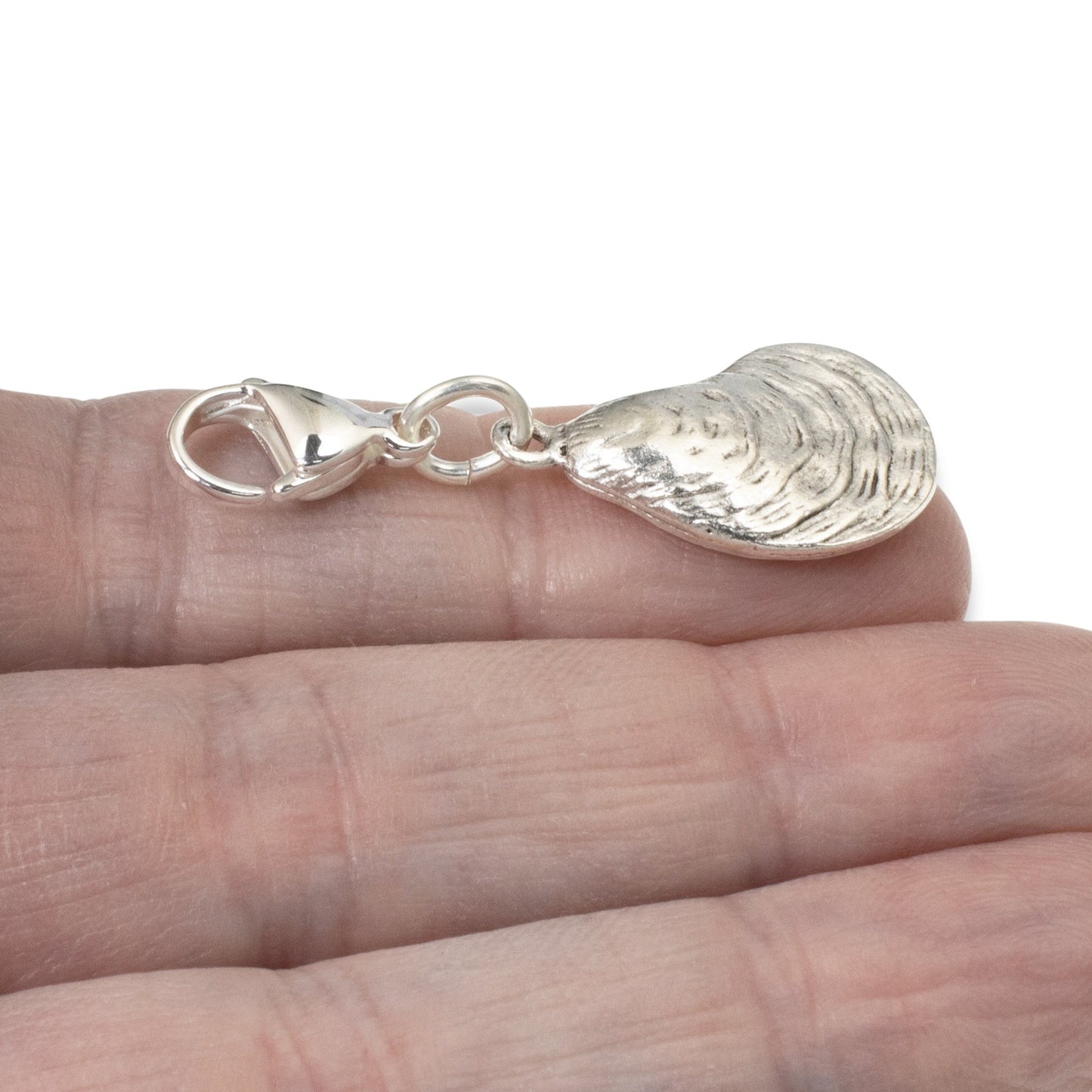 Hackberry Creek silver oyster shell charm with lobster clasp held on a hand, featuring detailed 3D coastal texture.