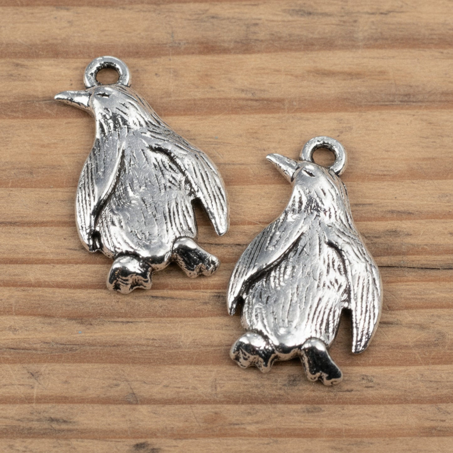 Hackberry Creek silver metal penguin charms with detailed feather texture for jewelry making on a wooden surface