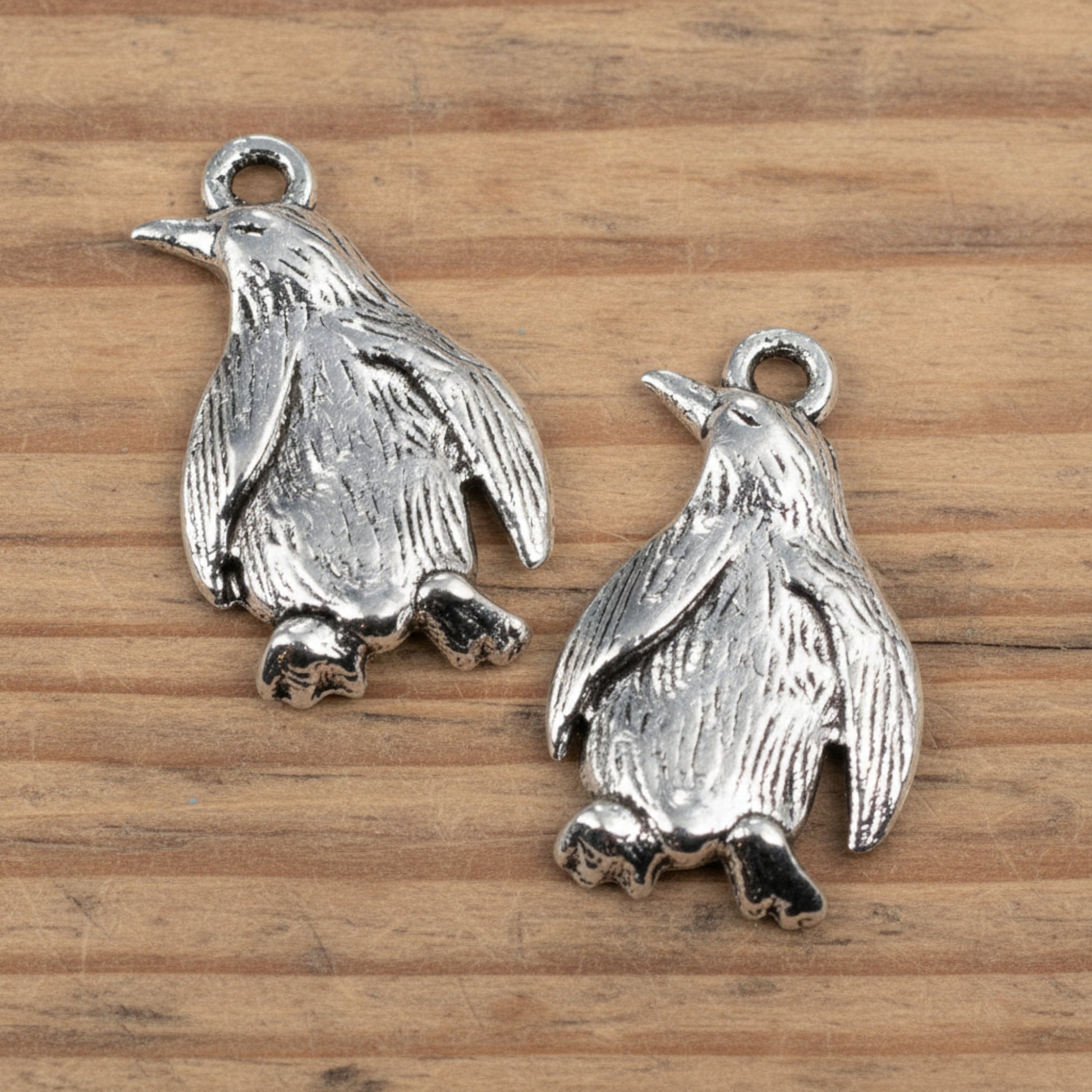 Hackberry Creek silver metal penguin charms with detailed feather texture for jewelry making on a wooden surface