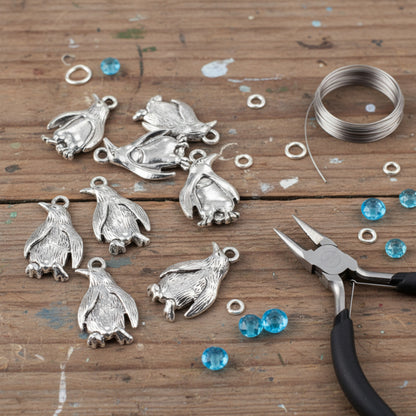 Hackberry Creek silver penguin charms with detailed feather texture, displayed with jewelry pliers and blue beads.