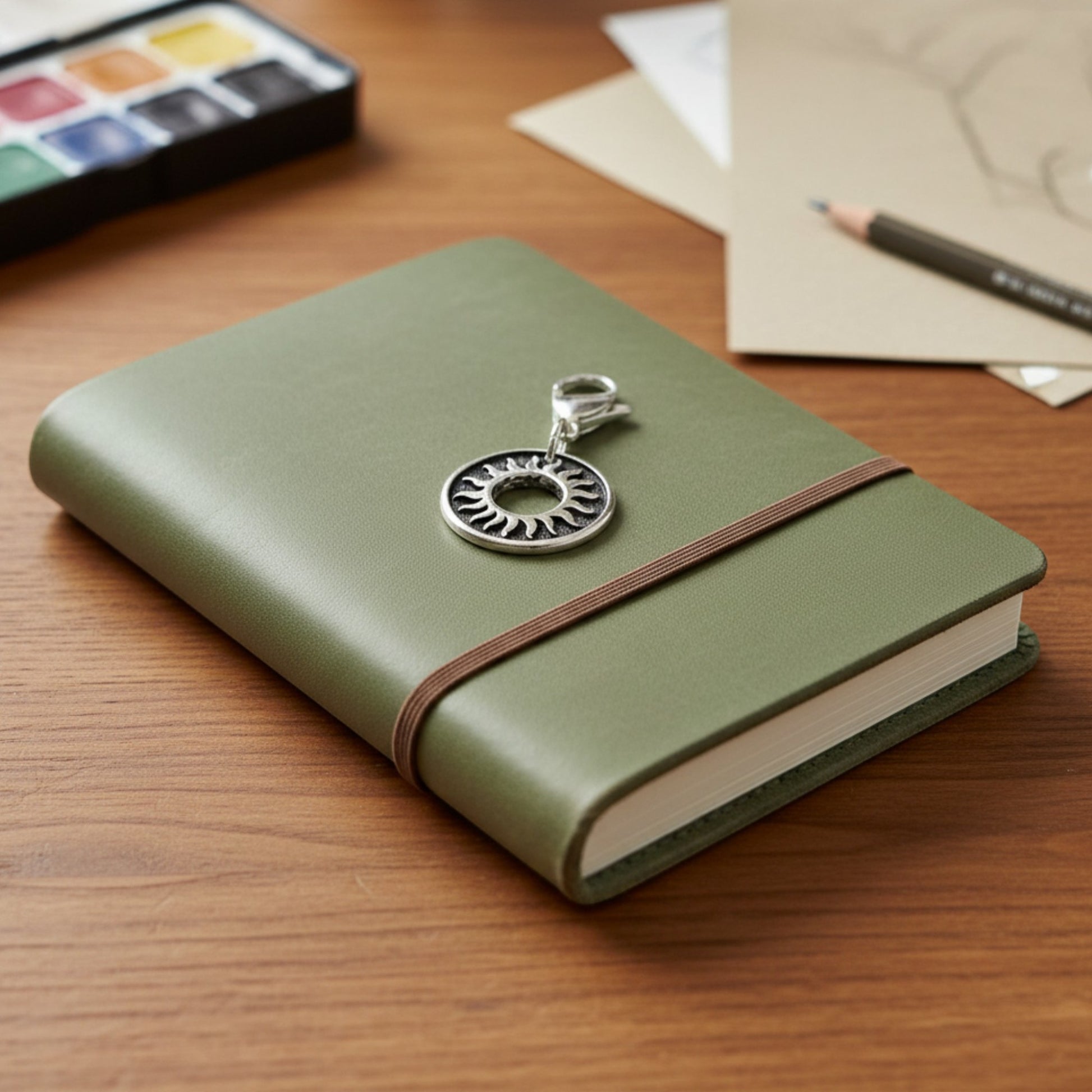 Hackberry Creek silver sun charm with lobster clasp attached to a green notebook on a wooden desk.