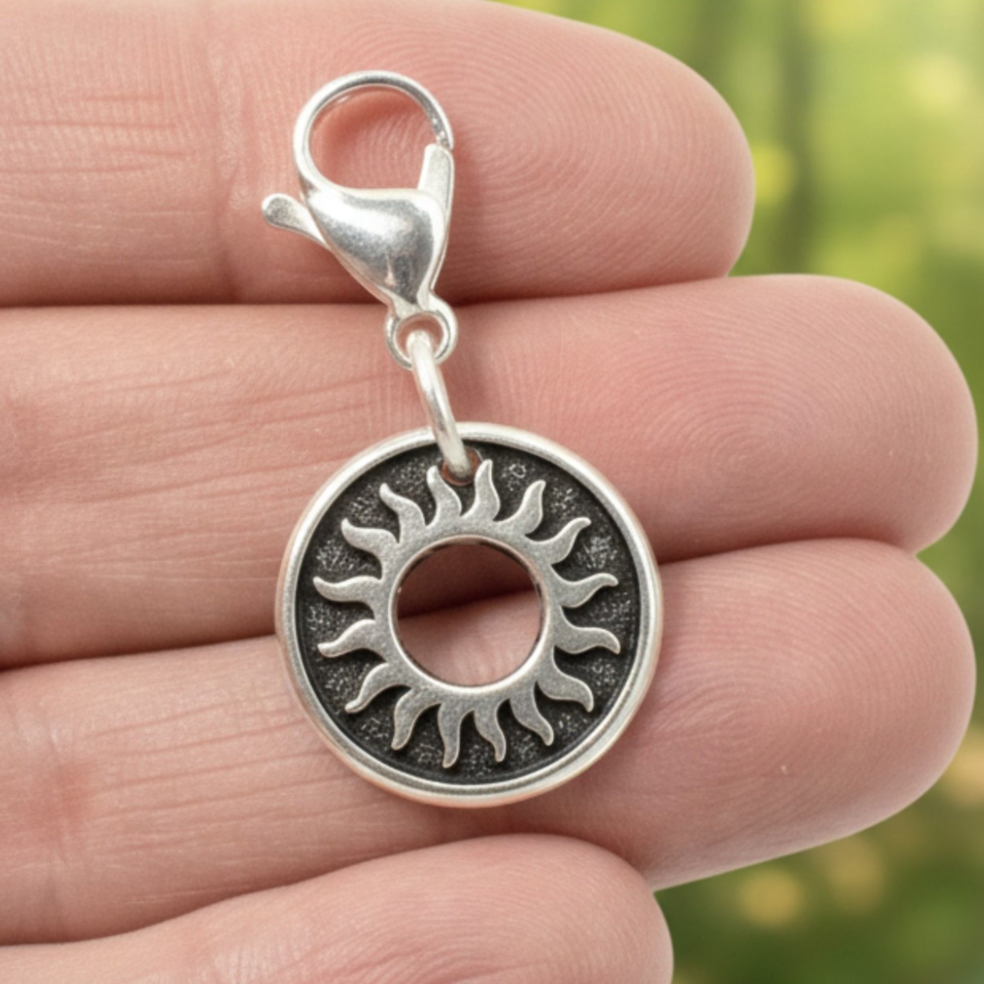 Hackberry Creek silver celestial sun charm with a stainless steel lobster clasp held in a hand against a forest background