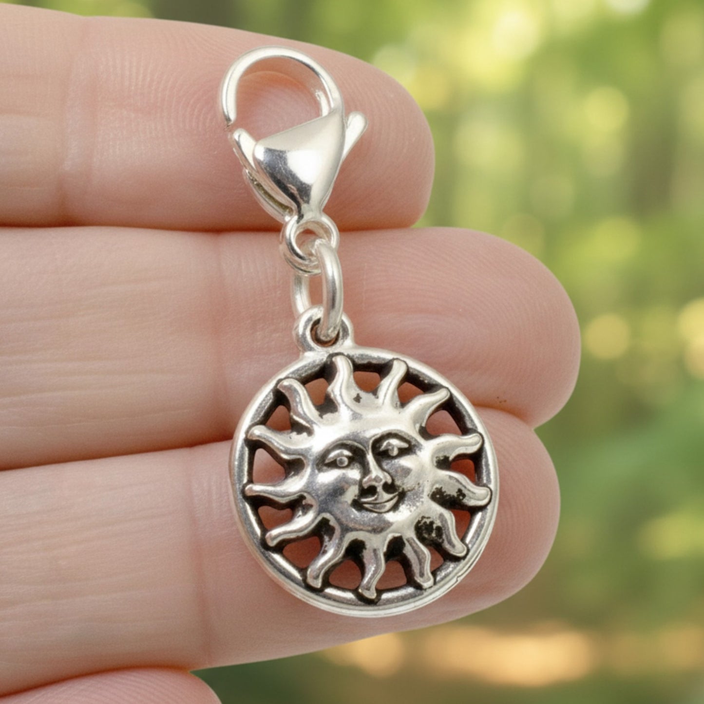 Hackberry Creek silver sun charm with a lobster clasp held between fingers against a blurred green forest background.