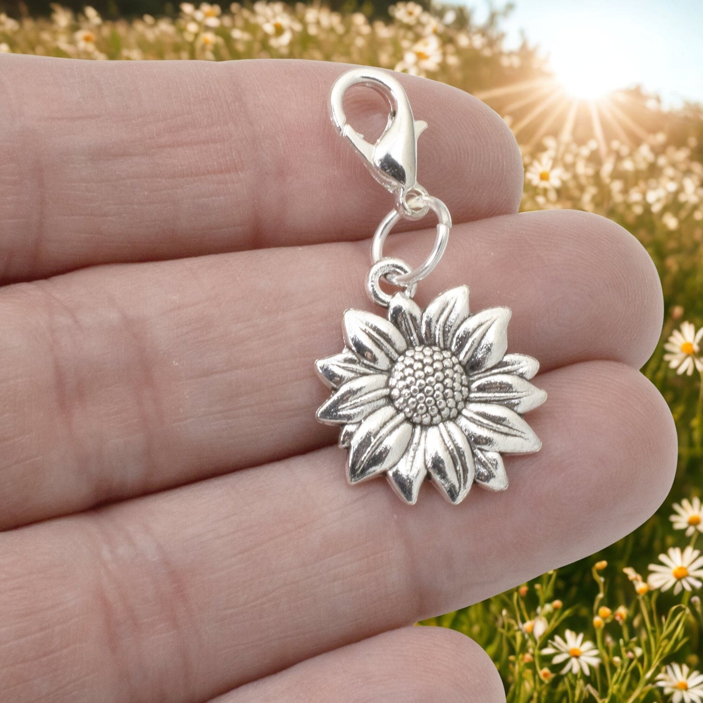 Hackberry Creek silver sunflower clip-on charm held in hand against a blurred field of daisies.