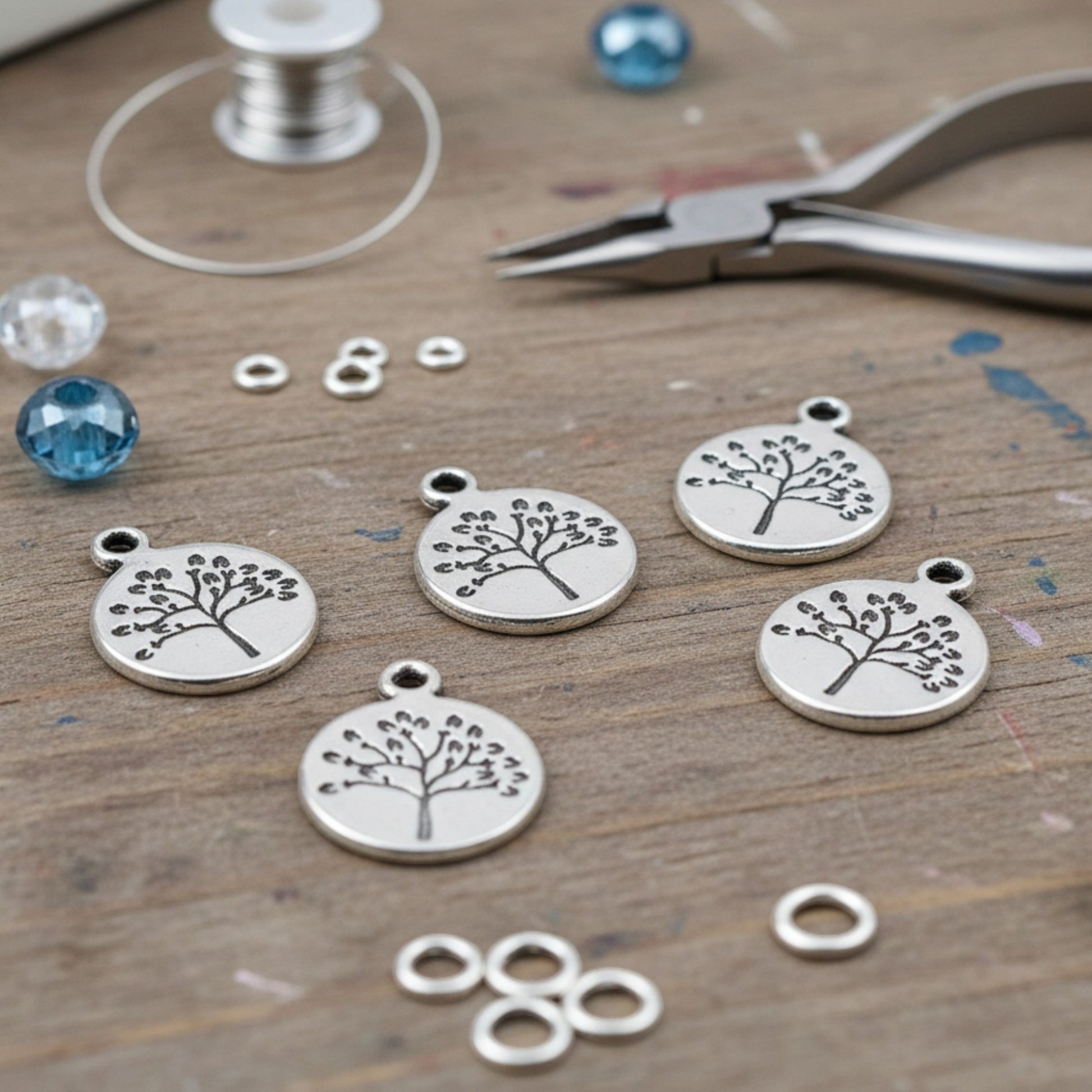 Hackberry Creek antique silver tree of life coin charms for jewelry making on a wooden craft table with tools